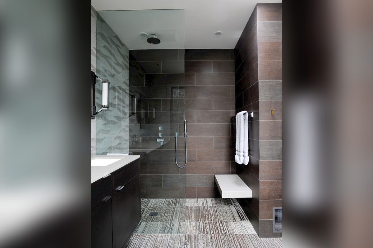 Modern bathroom with a walk-in shower, glass partition, wall-mounted showerhead, gray textured tiles, and a floating bench. A vanity with a mirror and folded towels is visible.