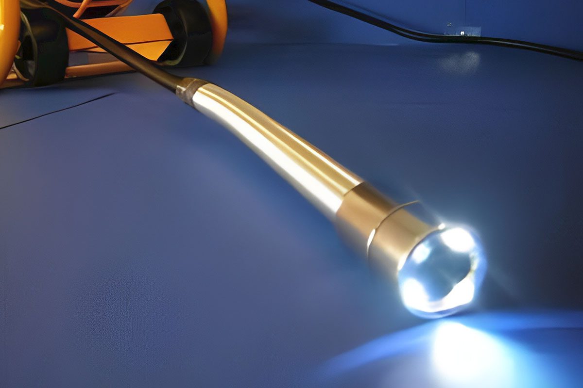 A close-up of a lit inspection camera with a bright LED light on a blue surface, connected to orange equipment.