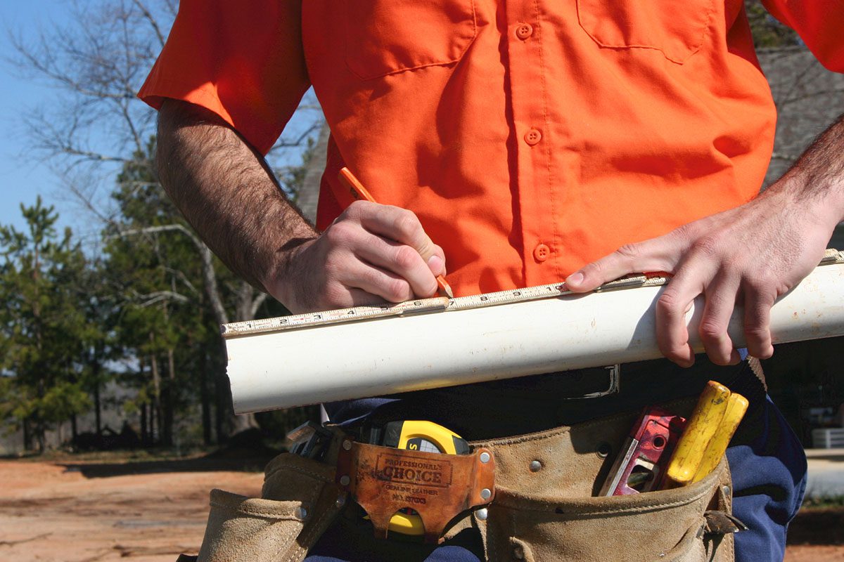 A person in an orange shirt and tool belt measures a white pipe with a tape measure in an outdoor setting.