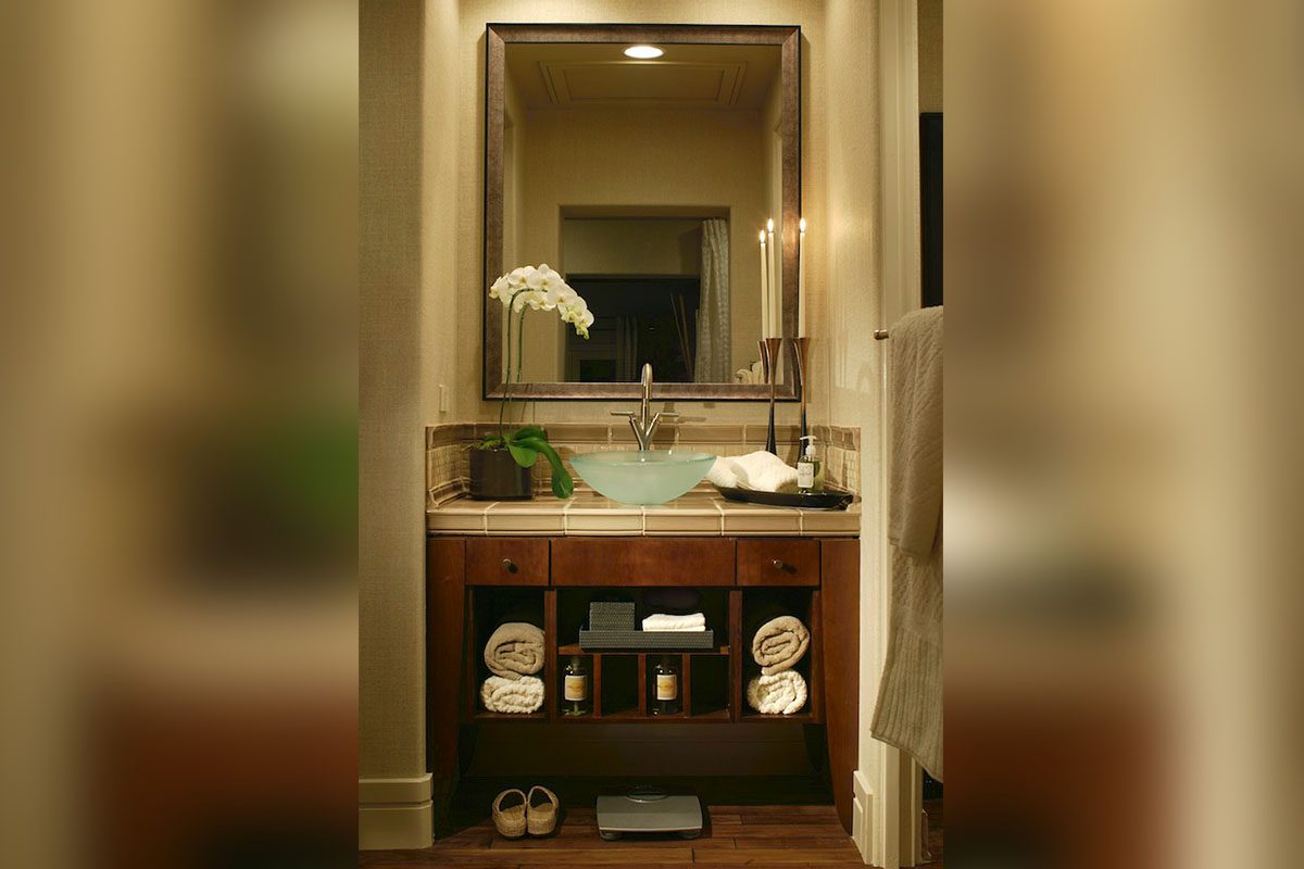 Bathroom vanity with a square mirror, glass bowl sink, and orchid. Towels and toiletries are on shelves below, with a digital scale on the floor. A plush robe hangs to the right.