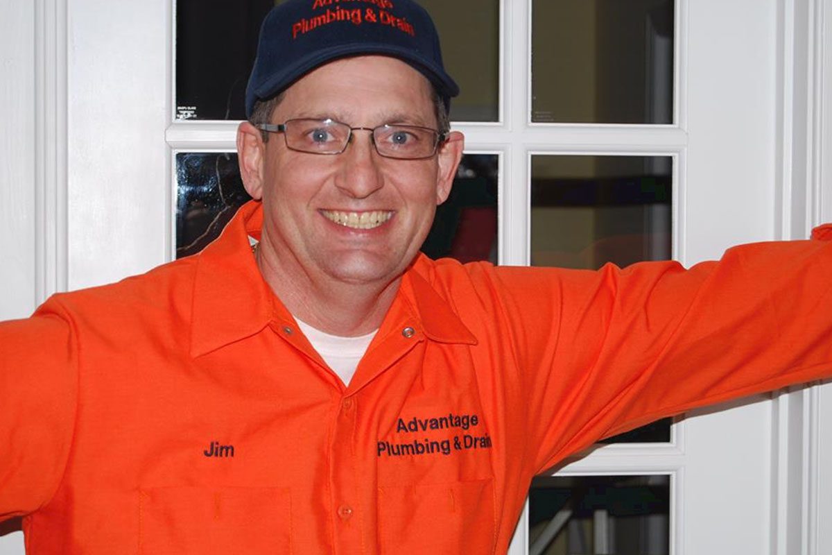 Man smiling, wearing glasses and an orange uniform with "Advantage Plumbing & Drain" text on the shirt and cap, standing in front of a windowed door.