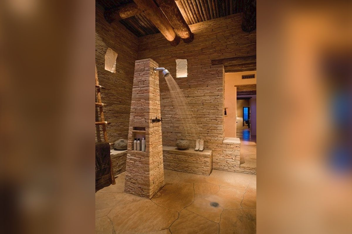 Rustic stone shower with a rain showerhead, wooden ceiling beams, and built-in shelving holding bath products. Warm lighting and stone tile flooring.