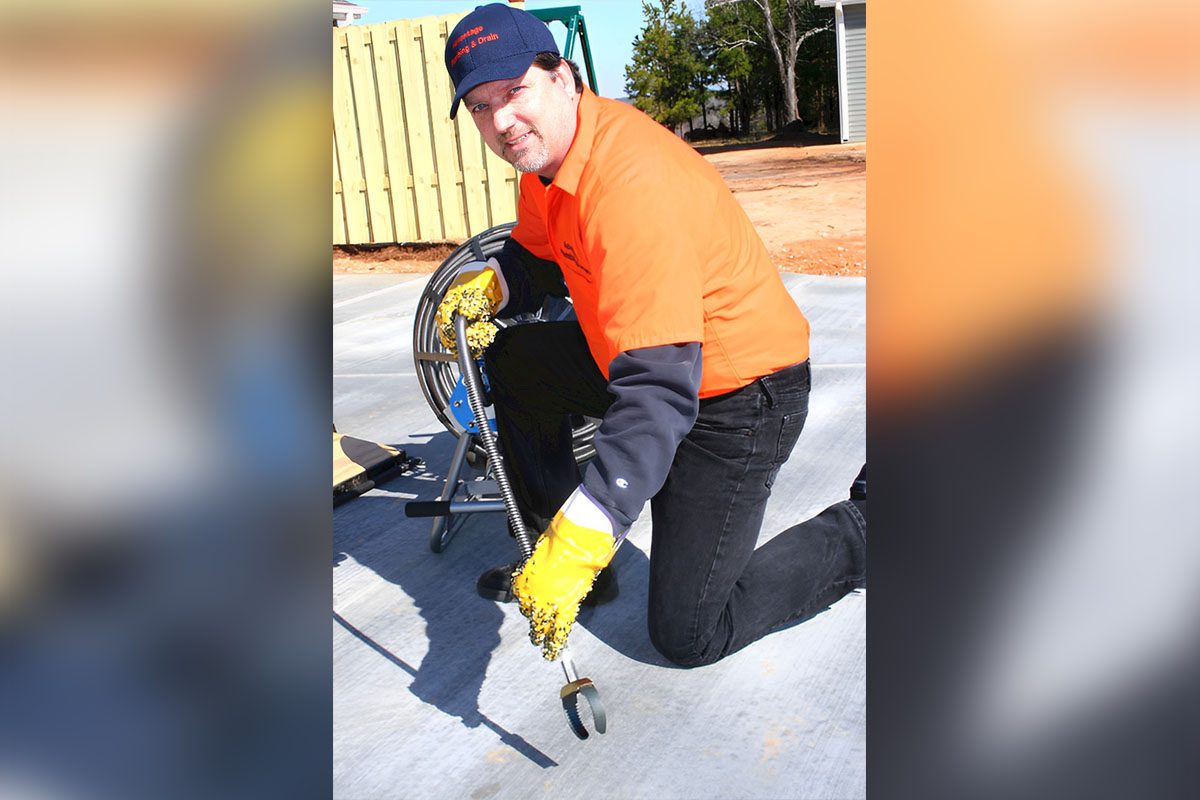 Person kneeling on concrete, wearing an orange shirt and cap, holding a plumbing snake with a camera. A reel with cable is in the background.