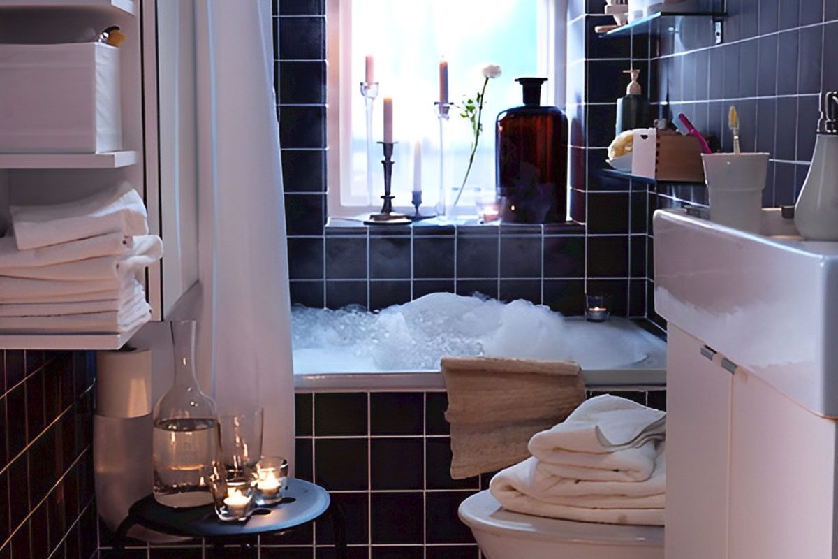 Cozy bathroom with a bubble-filled bathtub, candles, and folded towels. Dark tiled walls and a small sink are visible.