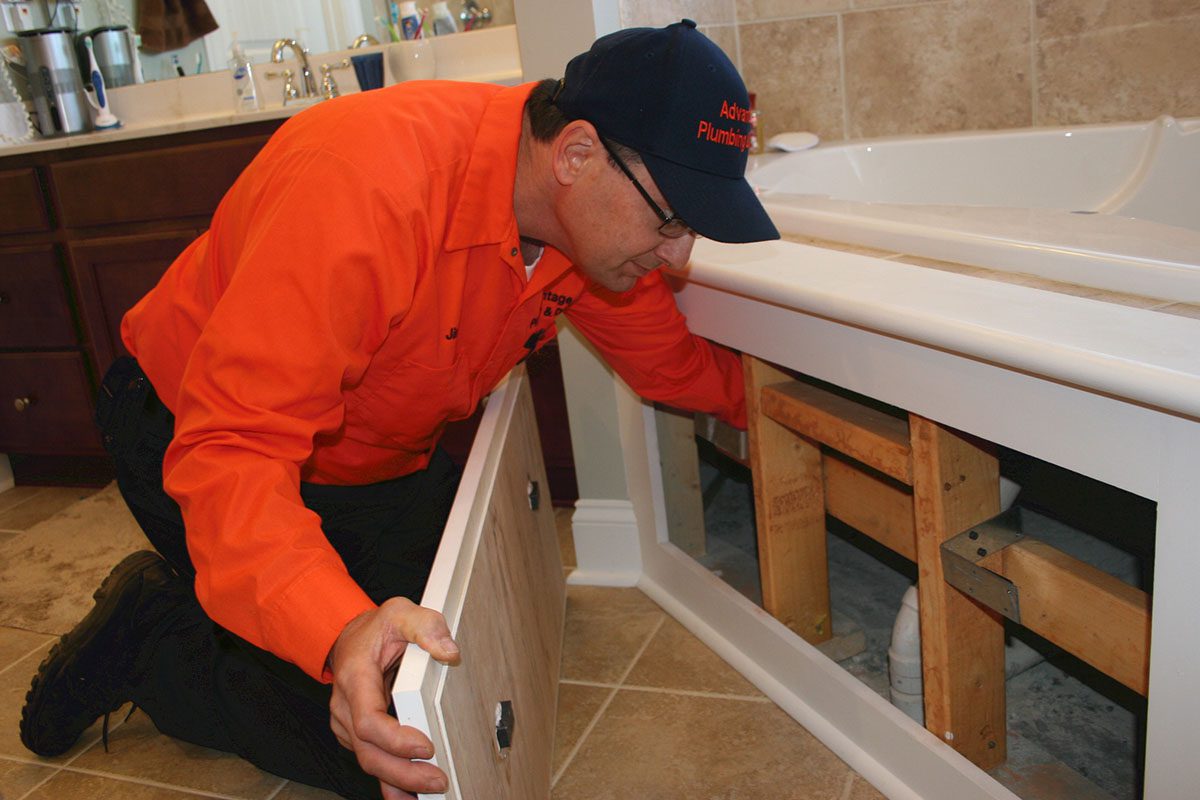 A plumber in an orange shirt and black cap examines the plumbing behind a bathtub panel in a bathroom.