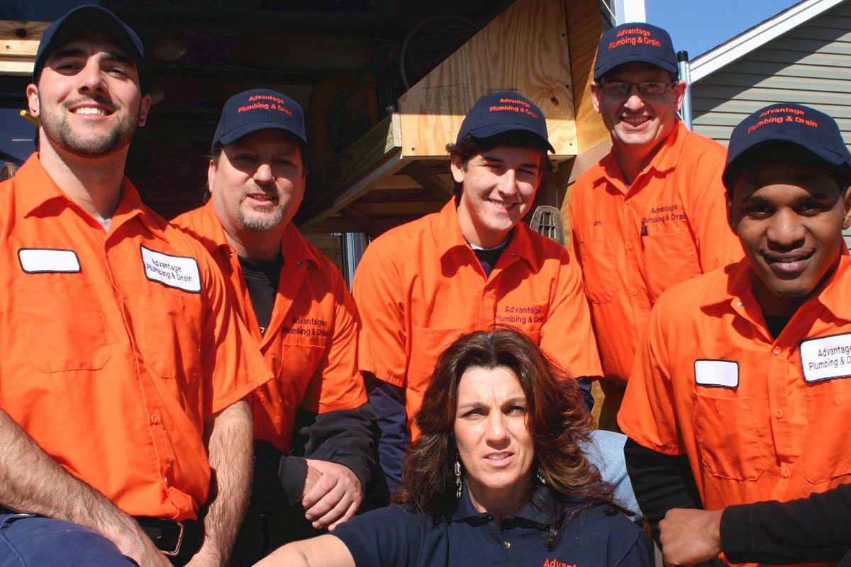 A group of five men in orange uniforms and one woman in a navy shirt pose outside. They are wearing caps with a company logo.