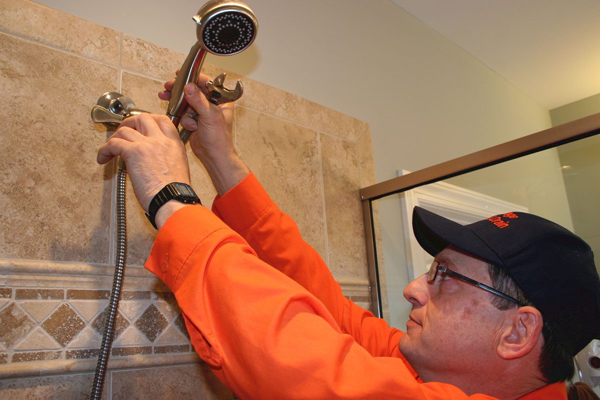 Person in an orange shirt and cap adjusts a showerhead in a tiled bathroom.