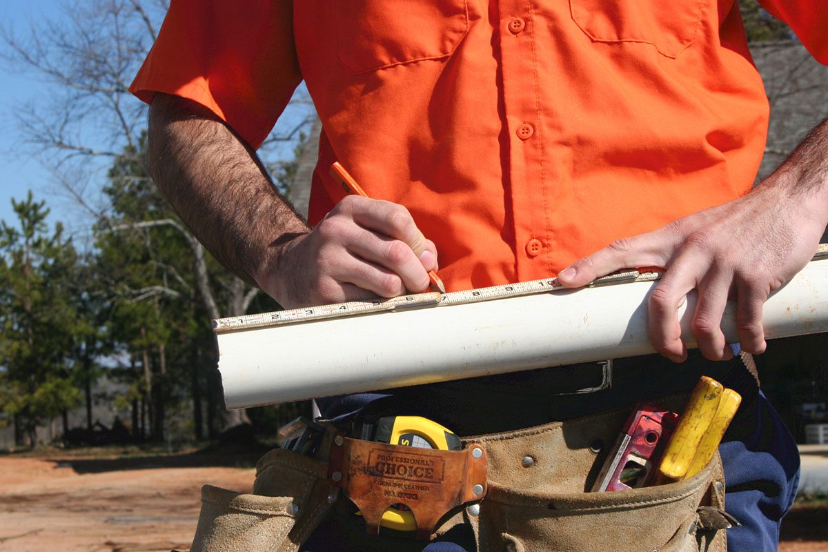 Person in an orange shirt using a tape measure and pencil to mark a white pipe, with a tool belt around their waist.