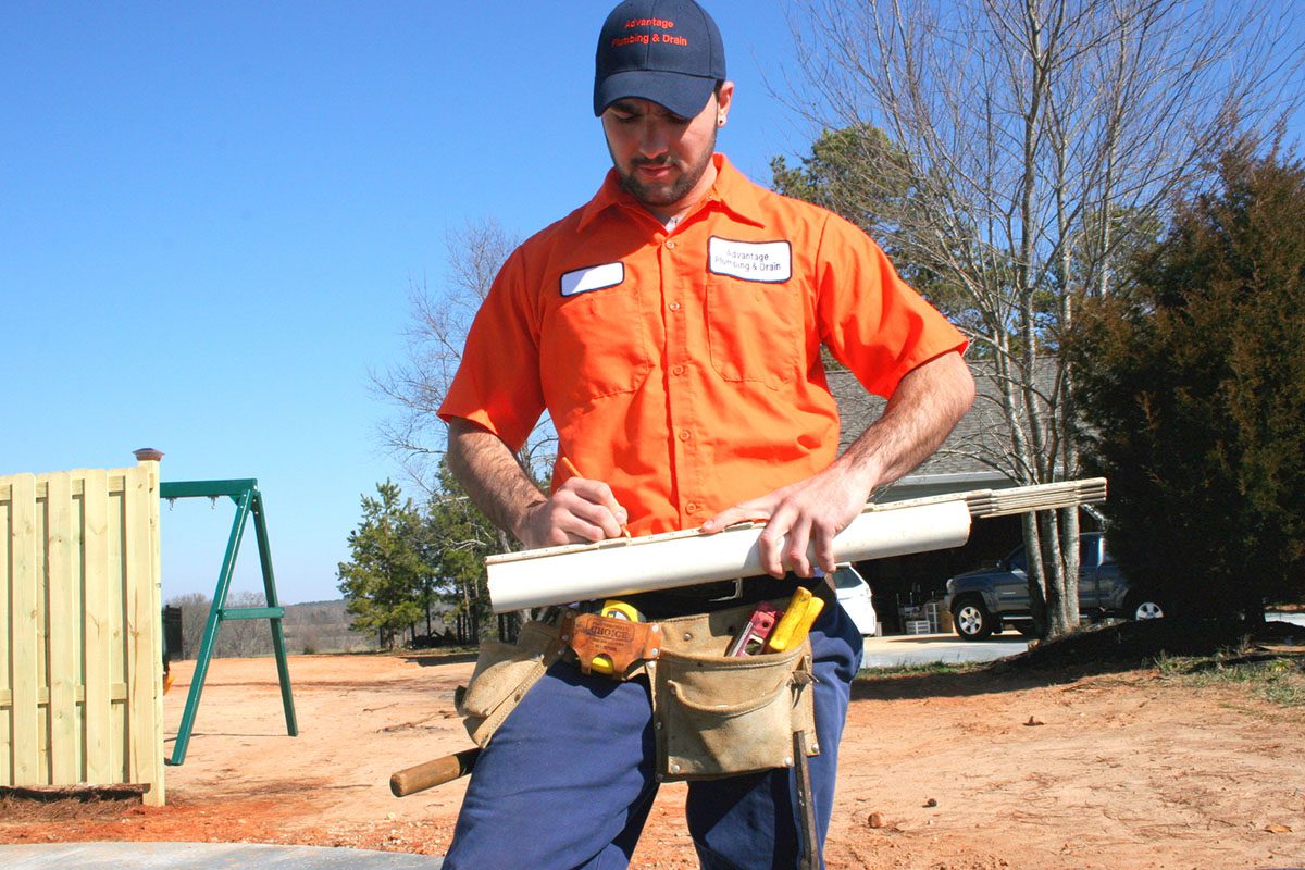 Worker in an orange shirt and blue pants takes notes on a rolled-up paper outdoors, wearing a tool belt.