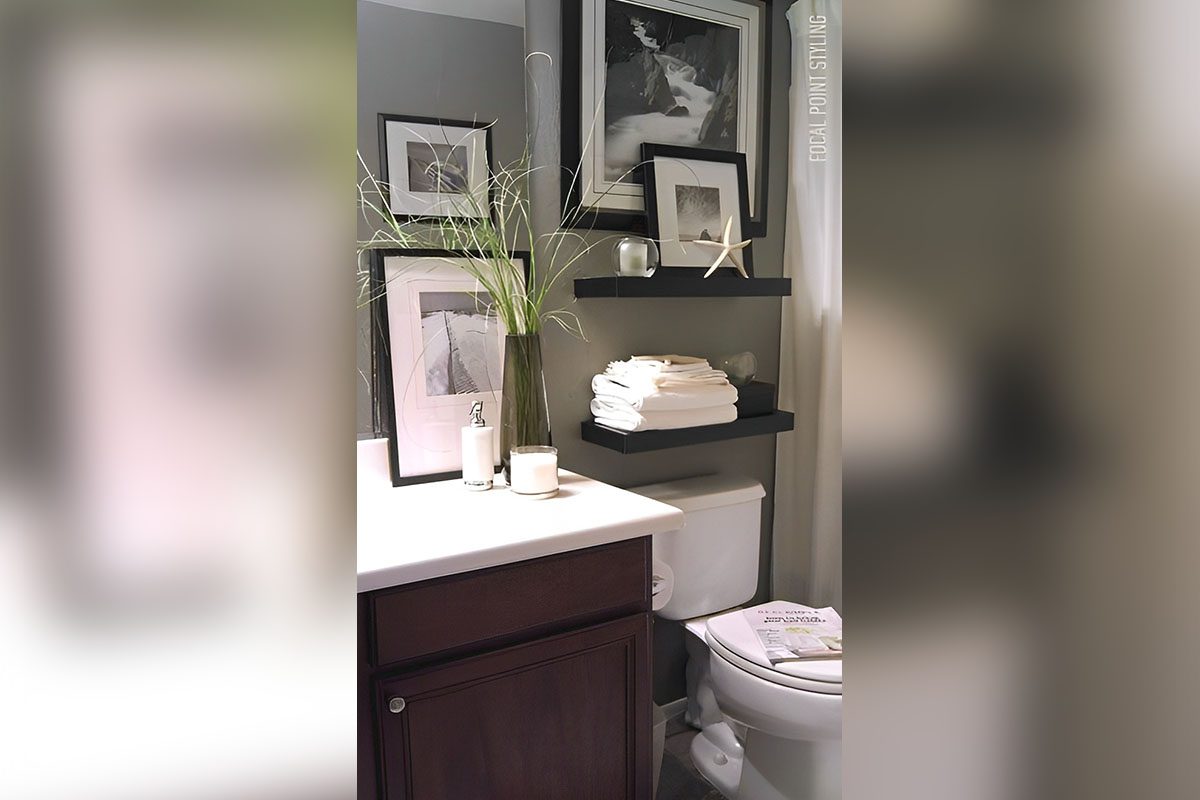Bathroom with framed black and white photos, a dark wooden cabinet, potted grass, a candle, and neatly stacked white towels beside a toilet.