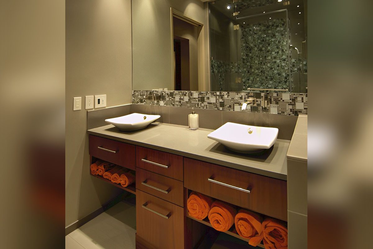 Modern bathroom with two square sinks on a countertop, large mirror, mosaic tile backsplash, and storage space below with neatly rolled orange towels.