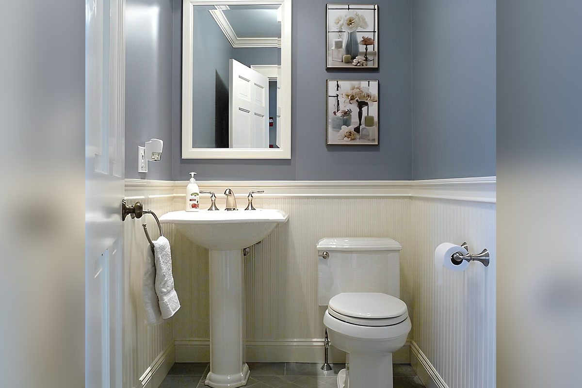 A bathroom with gray walls, a pedestal sink, a mirror, and framed pictures. There's a white toilet with a roll of toilet paper on the right and a towel on the left.