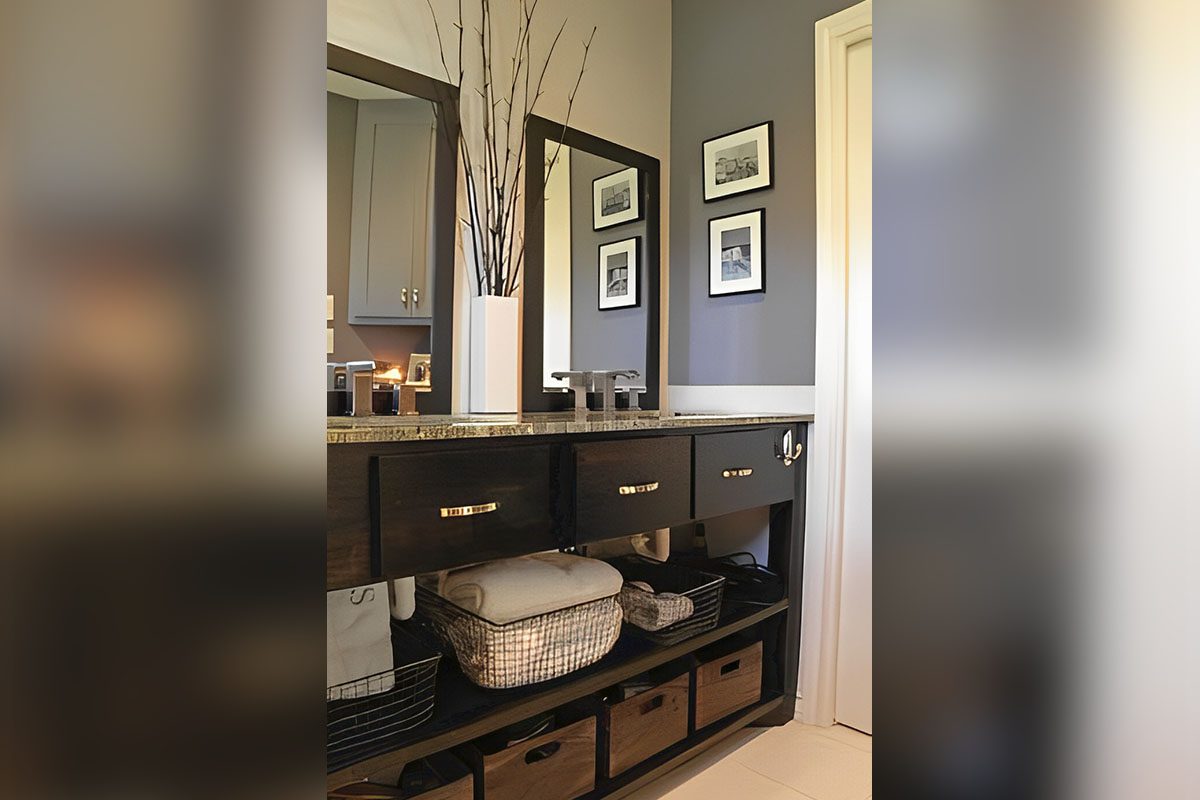 Modern bathroom with a dark wooden vanity, dual sinks, and a large mirror. Decorations include framed photos and a vase with branches. Storage baskets are neatly arranged below.
