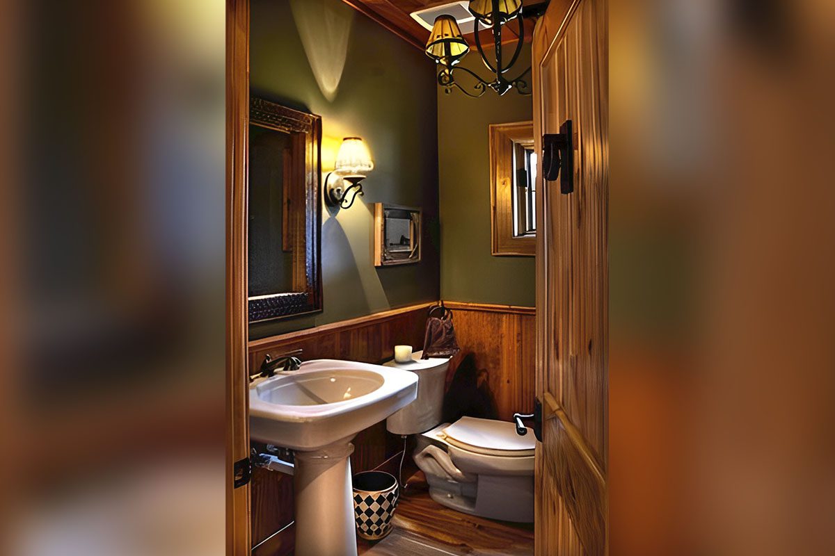 A small bathroom with wood paneling, a sink, mirror, and toilet. A chandelier hangs from the ceiling, and a wall sconce provides additional light.