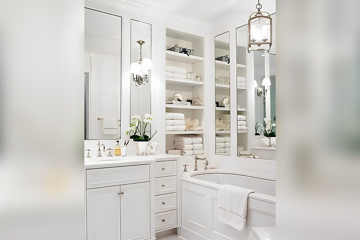 Elegant white bathroom with a bathtub, cabinets, and open shelves stocked with towels. Two large mirrors and a hanging light fixture are present. White orchids decorate the space.