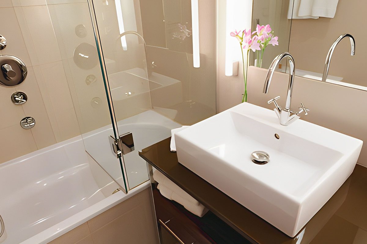 Modern bathroom with a square white sink on a dark countertop, chrome faucet, a glass-enclosed bathtub with showerhead, vertical lights, and a vase with pink flowers.