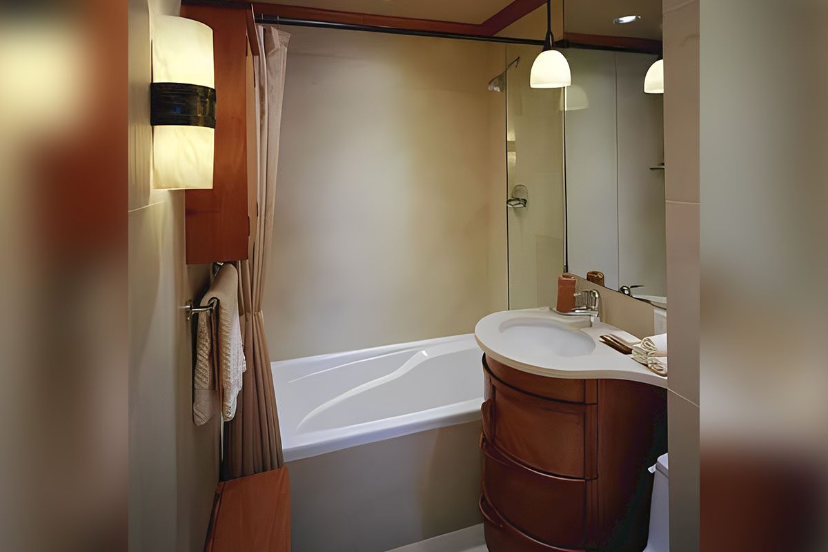 Small bathroom with a bathtub, curved wooden vanity, and a mirror. Two wall lights and a pendant light illuminate the space. A towel hangs on a rail beside the bath.