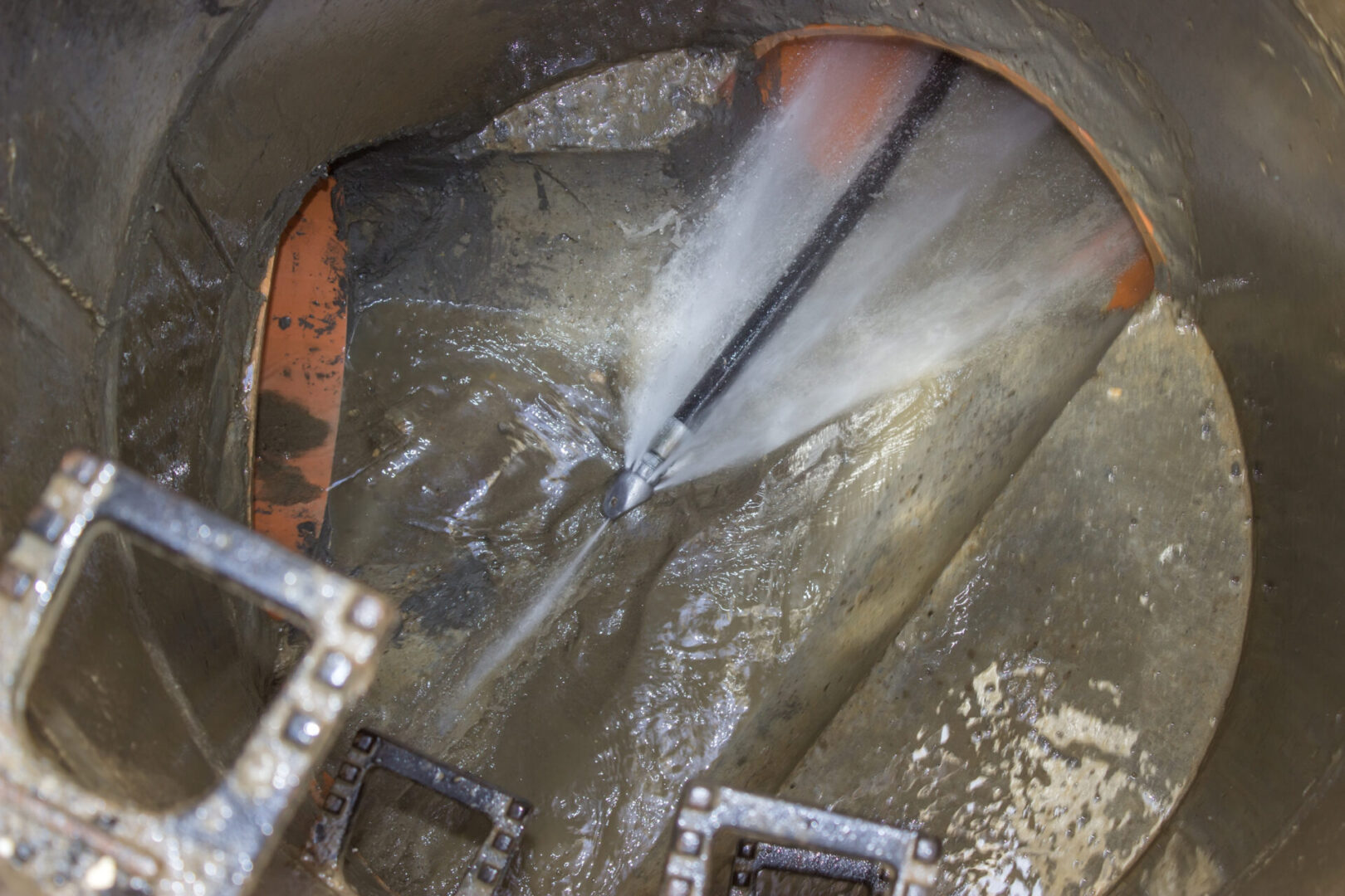 A sewer pipe being cleaned with high-pressure water, visible from inside manhole with metal rungs.