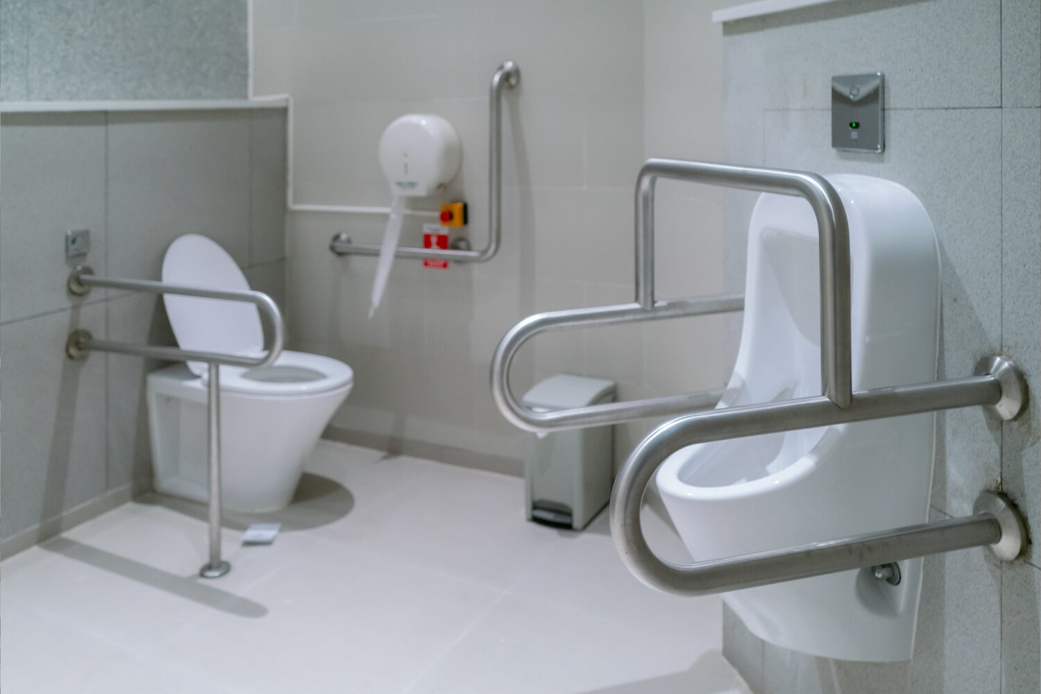An accessible restroom with a toilet and urinal, both equipped with stainless steel safety grab bars, and a wall-mounted hand dryer.
