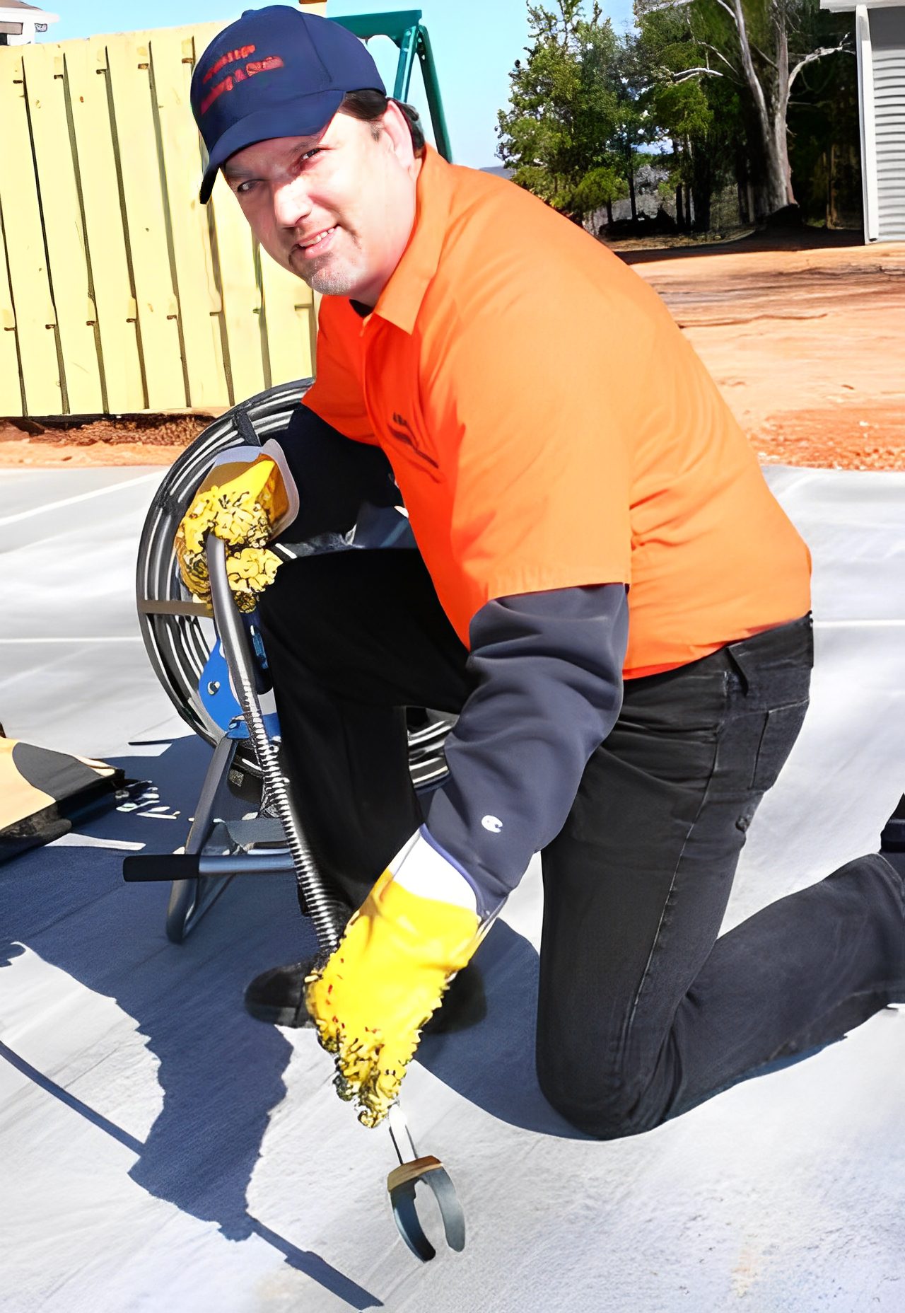 a0d0c688-95d9-4ec0-be34-1a0dbce55d3f A man wearing an orange shirt, black pants, and yellow gloves kneels on the ground while working with a coiled cable on a concrete surface.
