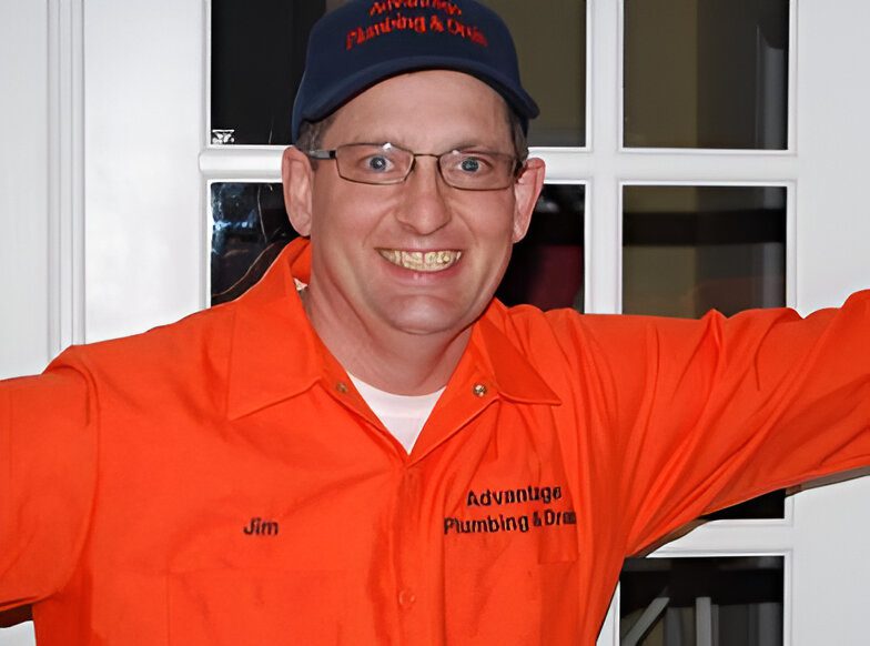 cea6090051eef5f518ca96337be504f5 A person smiling, wearing a navy cap and bright orange shirt with "Advantage Plumbing & Drain" and "Jim" embroidered on it, standing in front of a windowed door.